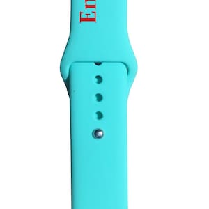 以下が含まれることがあります： 赤色の「Engraves Red」の文字が縦にプリントされた、ターコイズブルーの時計バンド。バンドにはピンと3つの調整穴があります。