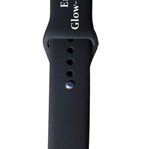 以下が含まれることがあります： 白文字で「Engraves Glow-in-the-Dark」と書かれた黒い時計バンド。バンドには調整用の穴と金属製の留め金が付いています。隅にある小さなボックスには「Activated Color」と書かれています。