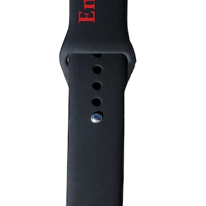 以下が含まれることがあります： 赤い文字で「Engraves Red」と書かれた黒い時計バンド。バンドは柔軟な素材でできており、調整用の穴があります。銀色のピンでバンドを固定します。