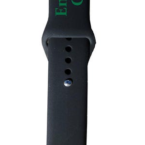 以下が含まれることがあります： 黒いシリコン製の時計バンド。緑色の文字で「Engraves Green」と書かれています。バンドはクラシックなデザインで、複数の調整穴とシルバー色のピンが付いています。