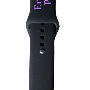 以下が含まれることがあります： 黒色の時計バンドで、ラベンダー色の「Engraves Purple」の文字が入っています。バンドはクラシックなデザインで、複数の調整穴とシルバー色のバックルが付いています。