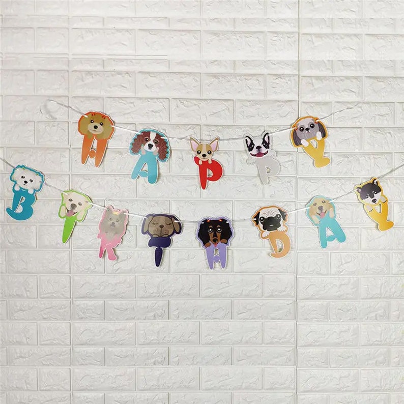 Puede incluir: Una pancarta de cumplea&ntilde;os feliz colorida con diferentes razas de perros en cada letra. La pancarta est&aacute; enhebrada en una cuerda blanca y est&aacute; colgada en una pared de ladrillo blanco.