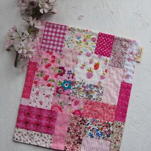 May include: A pink and white patchwork quilt with a variety of floral and geometric patterns. The quilt is made of squares and rectangles, and the seams are stitched with white thread. The quilt is on a white background.