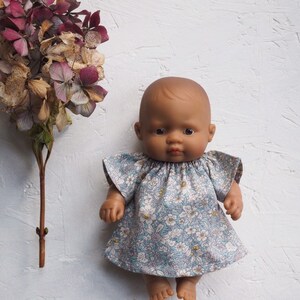 K&ouml;nnte beinhalten: Eine Puppe mit brauner Haut tr&auml;gt ein hellblaues Kleid mit Blumenmuster. Das Kleid hat kurze, ger&uuml;schte &Auml;rmel. Getrocknete Blumen in Lila- und Braunt&ouml;nen befinden sich auf der linken Seite des Bildes.
