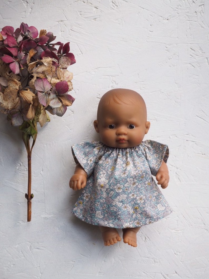 K&ouml;nnte beinhalten: Eine Puppe mit brauner Haut tr&auml;gt ein hellblaues Kleid mit Blumenmuster. Das Kleid hat kurze, ger&uuml;schte &Auml;rmel. Getrocknete Blumen in Lila- und Braunt&ouml;nen befinden sich auf der linken Seite des Bildes.