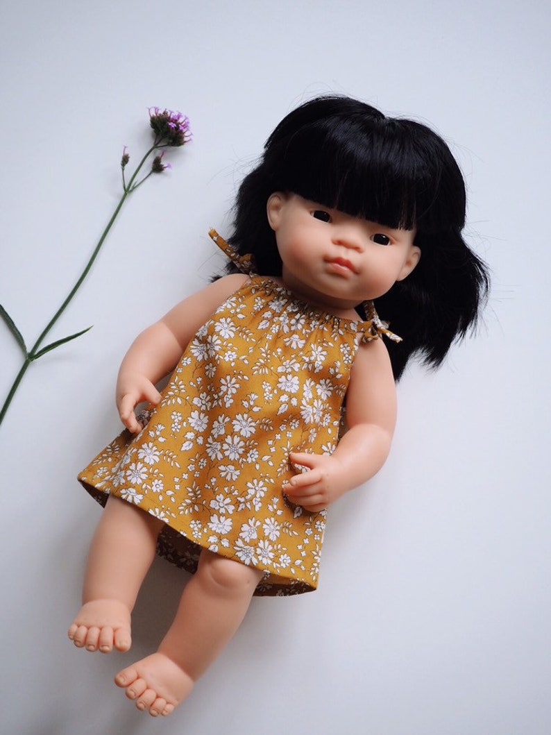 Op de afbeelding: Een pop met zwart haar die een gele bloemenjurk draagt. De pop heeft een lichte huidskleur en zit op een witte achtergrond.