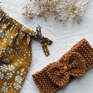 Op de afbeelding: Een bruin gebreid hoofdband met een strik. Het hoofdband ligt op een wit oppervlak met gedroogde bloemen op de achtergrond.