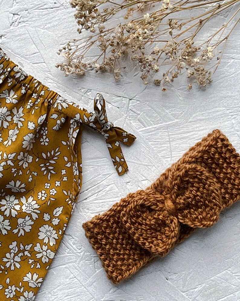 Op de afbeelding: Een bruin gebreid hoofdband met een strik. Het hoofdband ligt op een wit oppervlak met gedroogde bloemen op de achtergrond.