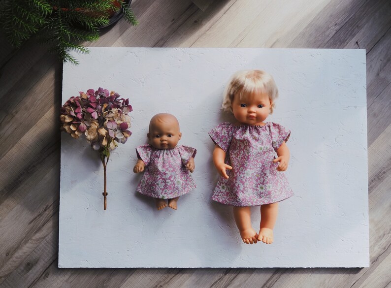 K&ouml;nnte beinhalten: Zwei Puppen, eine kleine und eine gr&ouml;&szlig;ere, werden auf einer wei&szlig;en Oberfl&auml;che gezeigt. Beide Puppen tragen passende Kleider mit einem Blumenmuster in Rosa- und Wei&szlig;t&ouml;nen. Links ist ein Trockenblumenarrangement positioniert.