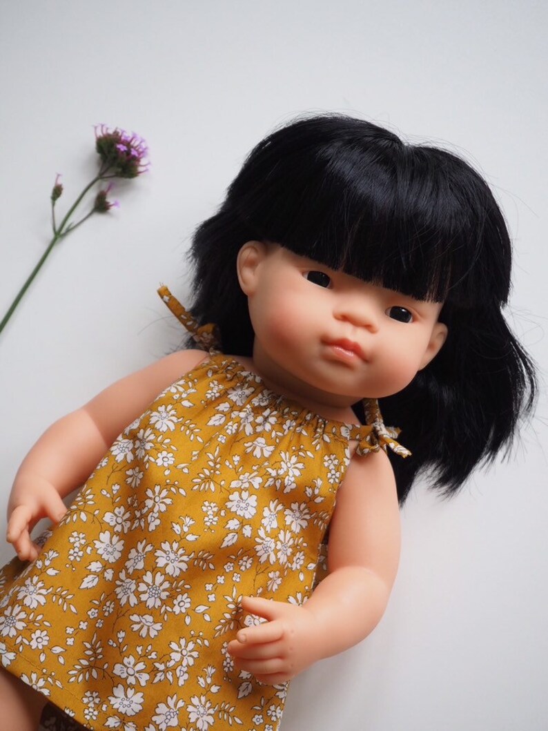 Op de afbeelding: Een pop met zwart haar en een gele bloemenjurk. De pop heeft een lichte huidskleur en bruine ogen.