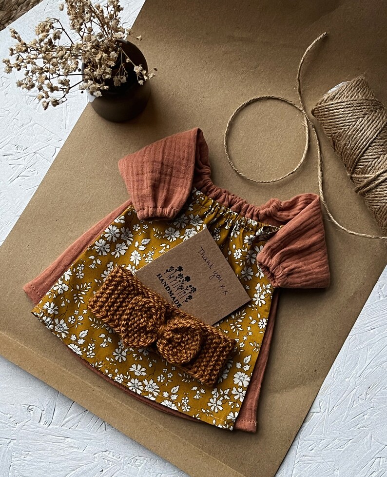 Op de afbeelding: Een bruine en gele bloemenjurk met een bruin hoofdband en een bedankkaartje met de tekst "Bedankt" en "Handgemaakt".