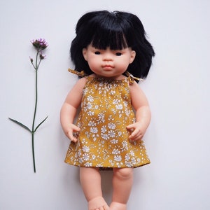 Op de afbeelding: Een pop met zwart haar en een gele bloemenjurk. De pop heeft een lichte huidskleur en draagt een gele jurk met witte bloemen.