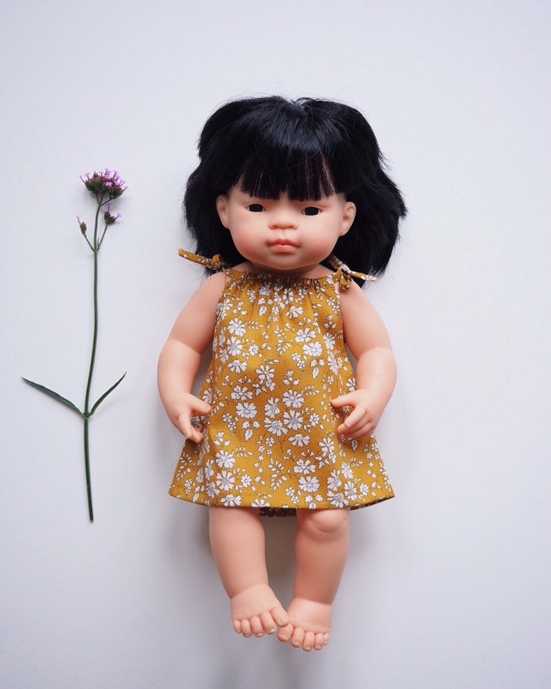 Op de afbeelding: Een pop met zwart haar en een gele bloemenjurk. De pop heeft een lichte huidskleur en draagt een gele jurk met witte bloemen.