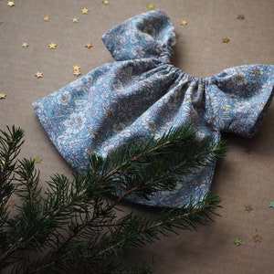 K&ouml;nnte beinhalten: Ein Puppenkleid mit blau-wei&szlig;em Blumenmuster. Das Kleid hat einen gerafften Ausschnitt und kurze &Auml;rmel. Der Text "Lalinka Doll Studio" ist sichtbar. Ein Zweig eines Nadelbaums und goldfarbene Sternkonfetti befinden sich im Vordergrund.