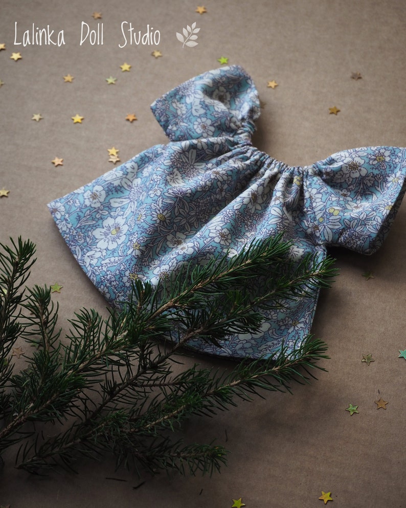 K&ouml;nnte beinhalten: Ein Puppenkleid mit blau-wei&szlig;em Blumenmuster. Das Kleid hat einen gerafften Ausschnitt und kurze &Auml;rmel. Der Text "Lalinka Doll Studio" ist sichtbar. Ein Zweig eines Nadelbaums und goldfarbene Sternkonfetti befinden sich im Vordergrund.