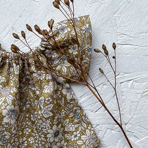 K&ouml;nnte beinhalten: Nahaufnahme eines Stoffes mit Blumenmuster. Der Stoff hat einen senfgelben Hintergrund mit wei&szlig;en und blauen Blumenmustern. Ein getrockneter Pflanzenstiel mit Samenkapseln befindet sich im Vordergrund und bildet einen nat&uuml;rlichen Kontrast.