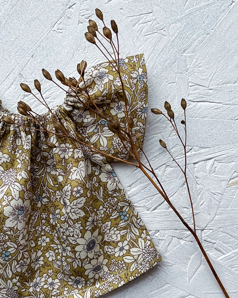 K&ouml;nnte beinhalten: Nahaufnahme eines Stoffes mit Blumenmuster. Der Stoff hat einen senfgelben Hintergrund mit wei&szlig;en und blauen Blumenmustern. Ein getrockneter Pflanzenstiel mit Samenkapseln befindet sich im Vordergrund und bildet einen nat&uuml;rlichen Kontrast.
