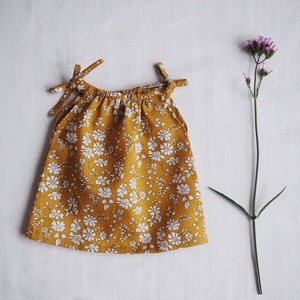 Op de afbeelding: Een gele, mouwloze jurk met een wit bloemenpatroon. De jurk heeft een geplooide halslijn en bandjes aan de schouders.