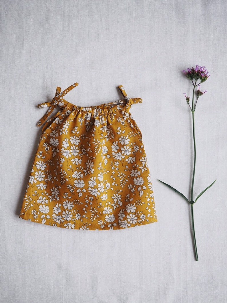 Op de afbeelding: Een gele, mouwloze jurk met een wit bloemenpatroon. De jurk heeft een geplooide halslijn en bandjes aan de schouders.