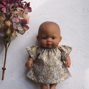 K&ouml;nnte beinhalten: Eine dunkelh&auml;utige Babypuppe in einem Kleid mit Blumenmuster und kurzen &Auml;rmeln. Das Kleid hat ein Muster aus wei&szlig;en und blauen Blumen auf gr&uuml;nem Grund. Die Puppe steht vor einem wei&szlig;en, strukturierten Hintergrund.