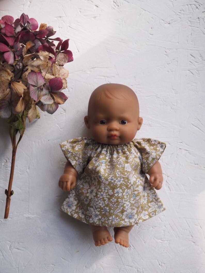 K&ouml;nnte beinhalten: Eine dunkelh&auml;utige Babypuppe in einem Kleid mit Blumenmuster und kurzen &Auml;rmeln. Das Kleid hat ein Muster aus wei&szlig;en und blauen Blumen auf gr&uuml;nem Grund. Die Puppe steht vor einem wei&szlig;en, strukturierten Hintergrund.