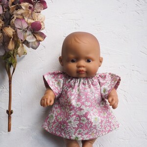 K&ouml;nnte beinhalten: Eine Puppe mit brauner Haut tr&auml;gt ein rosa Blumenkleid mit kurzen &Auml;rmeln. Das Kleid hat ein wei&szlig;es Blumenmuster. Getrocknete lila und braune Blumen befinden sich auf der linken Seite des Bildes.
