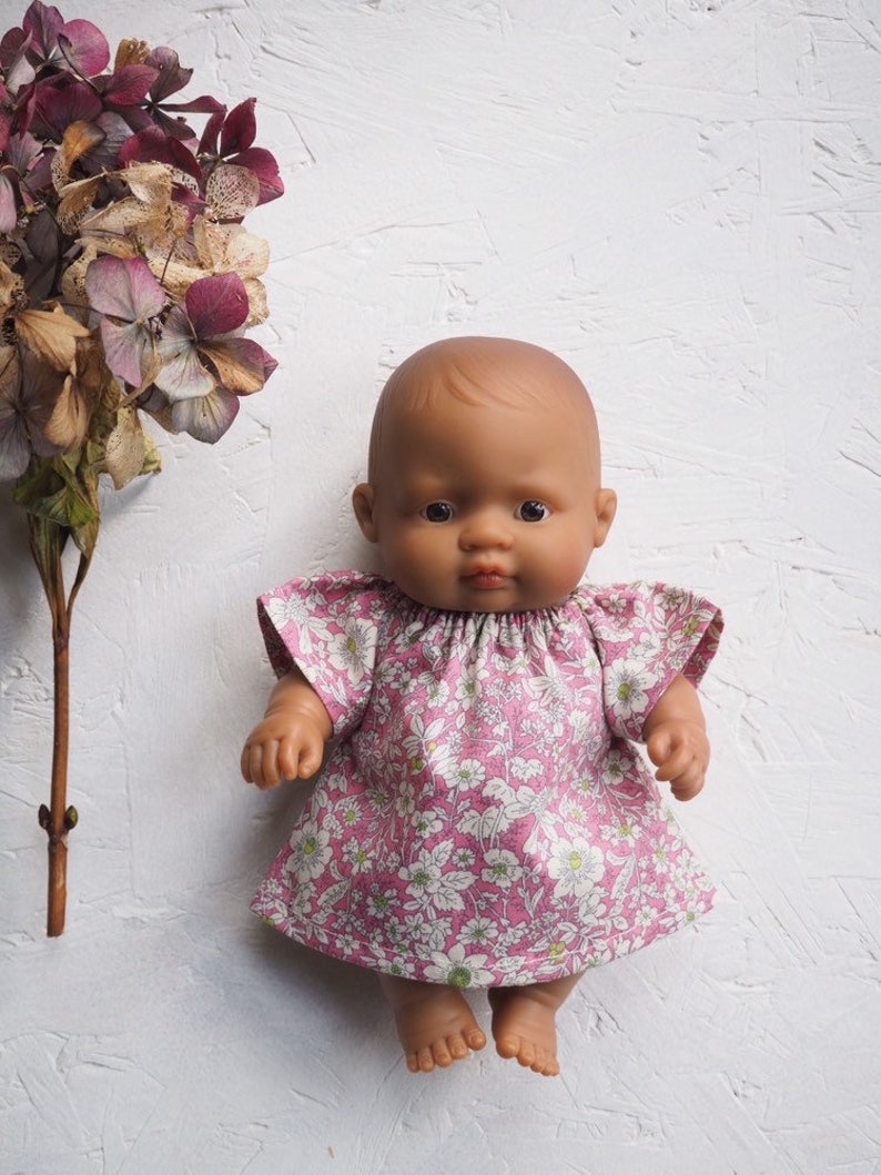 K&ouml;nnte beinhalten: Eine Puppe mit brauner Haut tr&auml;gt ein rosa Blumenkleid mit kurzen &Auml;rmeln. Das Kleid hat ein wei&szlig;es Blumenmuster. Getrocknete lila und braune Blumen befinden sich auf der linken Seite des Bildes.