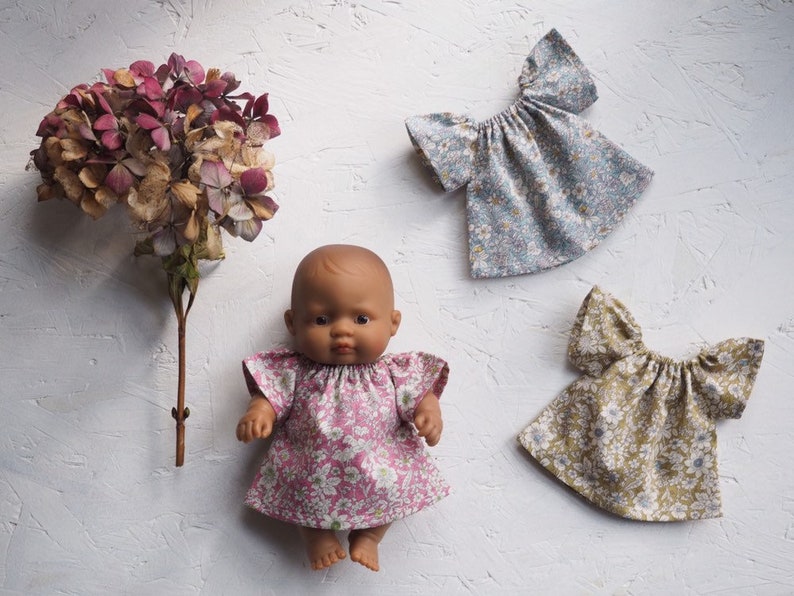 K&ouml;nnte beinhalten: Eine Puppe in einem rosa gebl&uuml;mten Kleid, mit passender Stoffpuppenkleidung in blau-gr&uuml;nen Blumenmustern. Links ist eine getrocknete Hortensienbl&uuml;te. Die Puppe hat braune Haut und braune Augen.