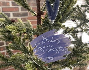 Puede incluir: Un adorno redondo transparente con un diseño de acuarela púrpura y el texto "Brahm's First Christmas 2023" en blanco.