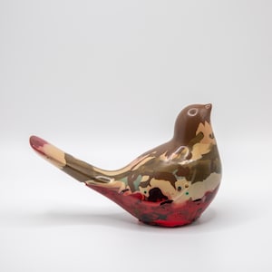 May include: A bird figurine featuring a smooth, glossy finish with a marbled pattern of brown, tan, olive green, and deep red colors. The sculpture has a rounded body, a pointed beak, and an elongated tail that extends upward and outward.