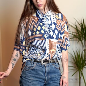 May include: A short-sleeved button-up shirt with a colourful geometric pattern in shades of blue, brown, white, and yellow. The shirt has a collared neckline and a button closure.