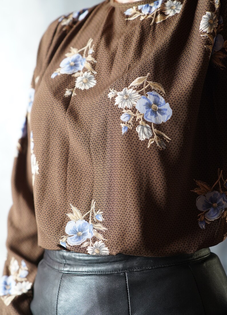 May include: A brown blouse with a floral print featuring blue and white flowers on a brown background with small white dots. The blouse has long sleeves and a round neckline. The blouse is paired with a black leather skirt.