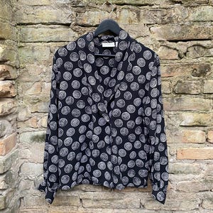 May include: A black and white long-sleeved button-up shirt with a circular pattern. The shirt is hanging on a black hanger against a brick wall.