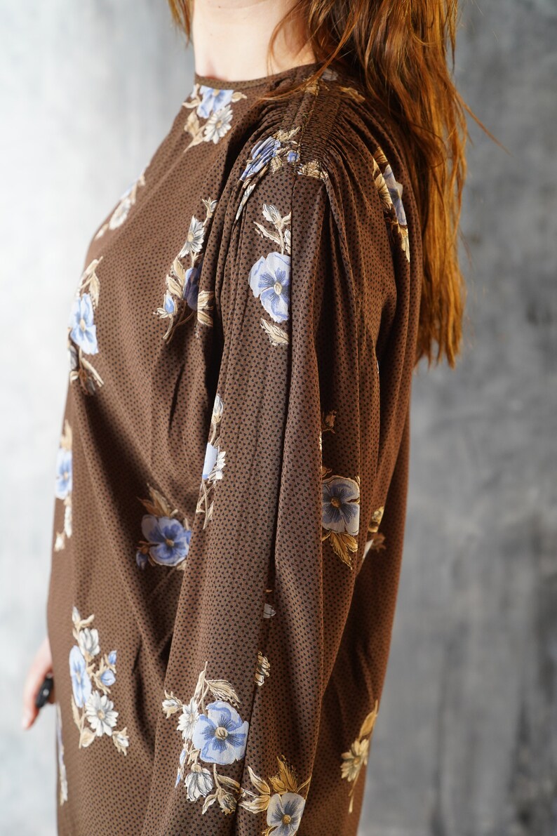 May include: A brown long-sleeved dress with a floral print featuring blue and white flowers. The dress has a gathered detail at the shoulders.