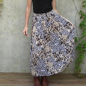 May include: A long, flowing skirt with a leopard print design in shades of brown, tan, and white. The skirt has a high waist and a full, gathered silhouette. The model is wearing a black top and brown boots.