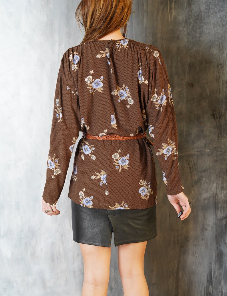 May include: A brown long-sleeved blouse with a floral print and a brown braided belt. The blouse has a round neckline and a relaxed fit. The model is wearing a black leather skirt.