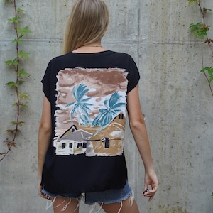 Könnte beinhalten: Schwarzes T-Shirt mit einem tropischen Landschaftsdruck auf der Rückseite, der Palmen und Gebäude in Braun-, Blau- und Weißtönen zeigt. Das T-Shirt hat kurze Ärmel und eine lockere Passform.