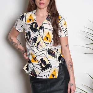 May include: A white wrap blouse with a short sleeve and a V-neck. The blouse has a yellow, black, and purple abstract print with a geometric design.