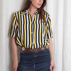 Pode incluir: Uma camisa de manga curta com listras verticais em amarelo, branco e azul marinho. A camisa tem gola e botões dourados. Um cinto de couro marrom com fivelas decorativas prateadas é usado com uma saia jeans.