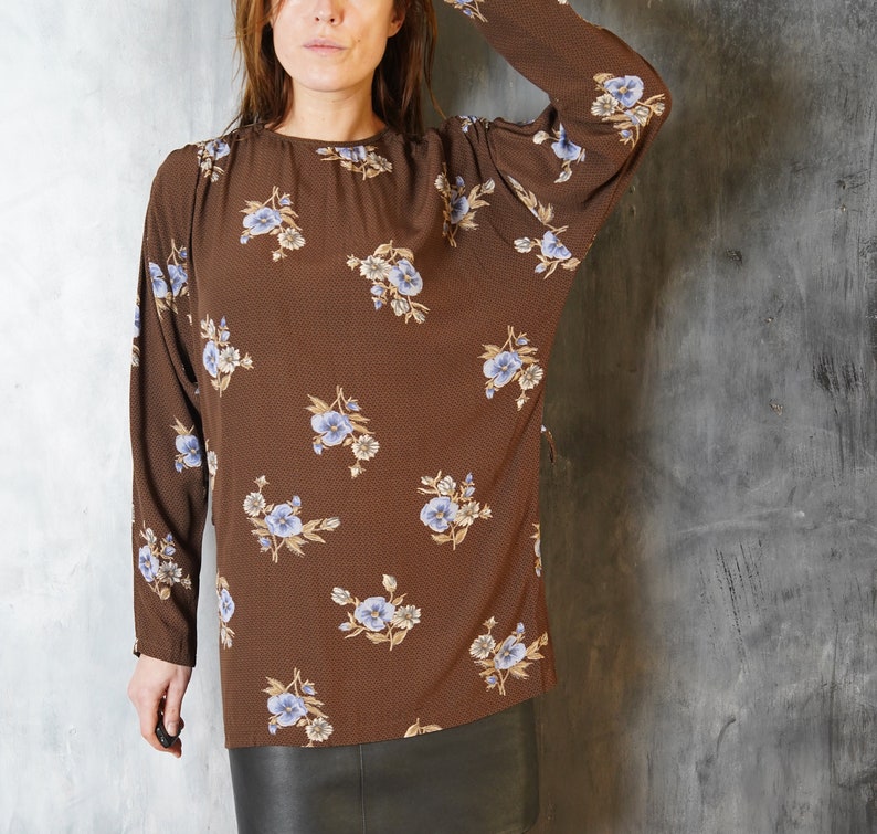 May include: A brown long-sleeved blouse with a floral print featuring blue and white flowers. The blouse has a round neckline and a relaxed fit.