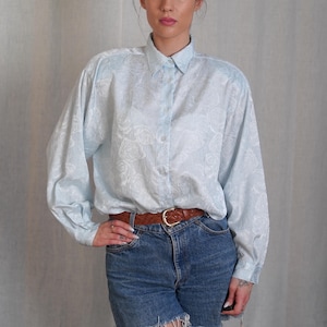 May include: A light blue button-down shirt with a white floral pattern. The shirt has long sleeves and a collared neckline. The model is wearing a brown leather belt.