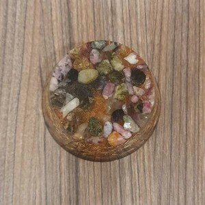 Pu&ograve; includere: Un sottobicchiere rotondo in resina con una variet&agrave; di pietre colorate incastonate. Le pietre sono principalmente verdi, rosa e marroni e sono incastonate in una resina trasparente.