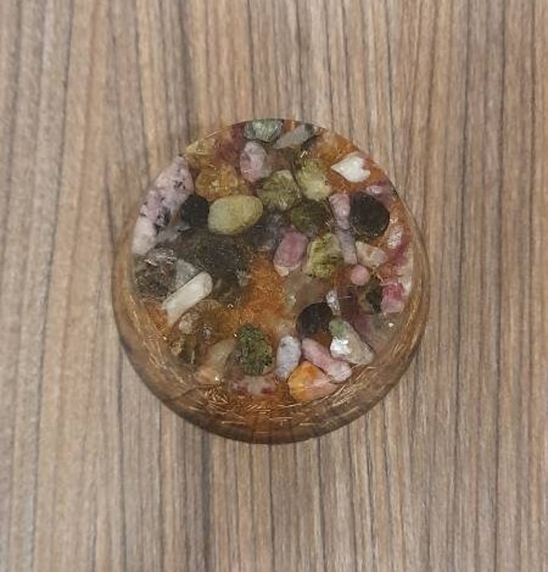 Pu&ograve; includere: Un sottobicchiere rotondo in resina con una variet&agrave; di pietre colorate incastonate. Le pietre sono principalmente verdi, rosa e marroni e sono incastonate in una resina trasparente.