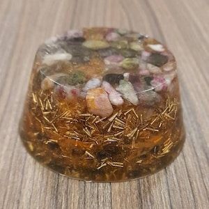 Pu&ograve; includere: Una sfera rotonda in resina trasparente riempita di brillantini dorati e cristalli di vari colori. I cristalli sono rosa, verdi e neri, e la sfera &egrave; appoggiata su una superficie di legno.