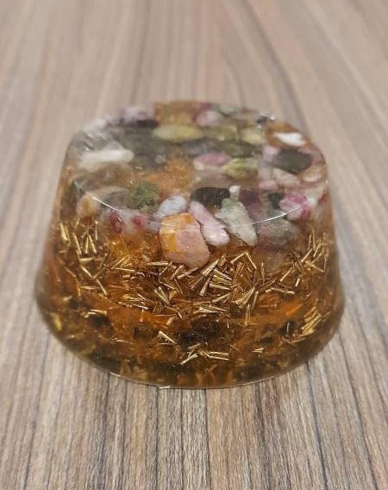 Pu&ograve; includere: Una sfera rotonda in resina trasparente riempita di brillantini dorati e cristalli di vari colori. I cristalli sono rosa, verdi e neri, e la sfera &egrave; appoggiata su una superficie di legno.