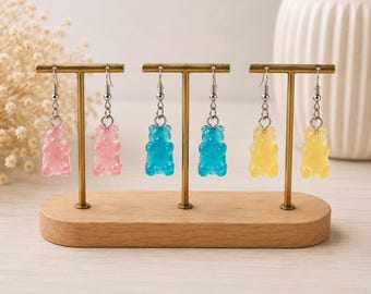 Könnte beinhalten: Drei Paar Gummibärchen-Ohrringe hängen an einem goldfarbenen T-förmigen Metallständer. Die Ohrringe bestehen aus durchscheinenden rosa, blauen und gelben bärenförmigen Anhängern, die an silberfarbenen Metall-Ohrhaken mit kleinen Ringen befestigt sind.