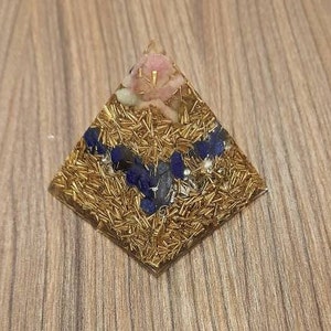 Può includere: Piramide dorata con cristalli blu e rosa incorporati nella resina. La piramide è su una superficie di legno.