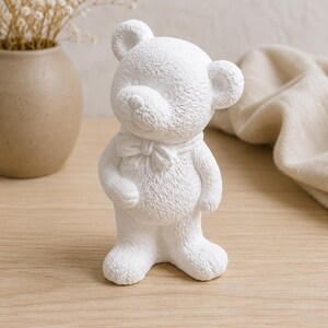 Può includere: Una statuetta di orso bianca e testurizzata con un fiocco scolpito attorno al collo. L'orso presenta una testa arrotondata, piccole orecchie e una finitura superficiale granulosa che imita l'aspetto di una pelliccia morbida, in piedi.