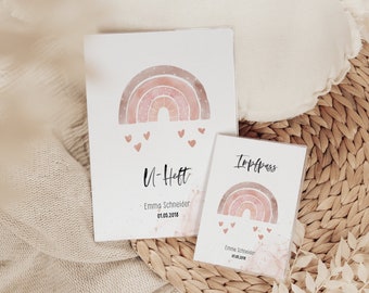 Op de afbeelding: Twee witte notitieboekjes met een aquarel regenboogontwerp in roze en perziktinten, met kleine roze hartjes eronder. Het grotere notitieboekje heeft de tekst "U-Heft", de naam "Emma Schneider" en de datum "01.05.2018". Het kleinere notitieboekje zegt "Impfpass".