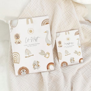 May include: Two white photo albums with a light brown border. The larger album has the text "U-H" followed by "LENI SCHNEYDERN 18.06.2023". The smaller album has the text "Lenipfags" followed by "LENI SCHNEYDERN 18.06.2023". Both albums have a white background with illustrations of a sun, a rainbow, a sailboat, a bird, a goose, a bear, an elephant, and a lion.